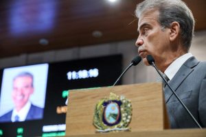 Deputado Coronel Alberto Feitosa, usando terno cinza, fala na tribuna, enquanto uma foto dele no telão do Plenário aparece ao fundo