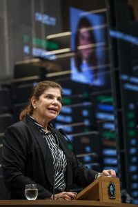 Deputada Socorro Pimental fala na tribuna, usando blazer preto sobre um camisa preta e branca. Ao fundo, reflexo do telão com o resto dela sobre o vidro das galerias