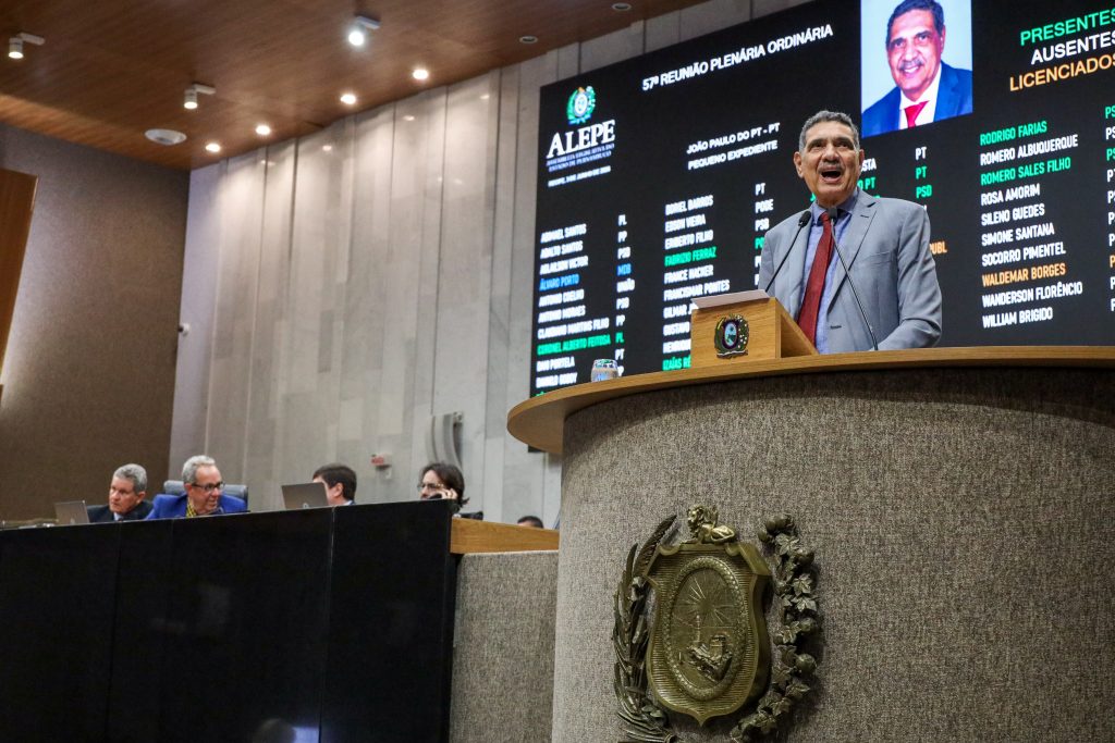 Deputado João Paulo aparece na parte superior direita num plano aberto na tribuna, com a Mesa Diretora ao fundo na esquerda, e telão com painel dos deputados ao fundo
