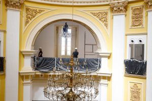 Detalhe dos frisos nos arcos internos do Palácio Joaquim Nabuco, nas cores branca, amarela e dourada. Ao centro, o lustre pendente em dourado. As grades seguem cobertas por lonas pretas.