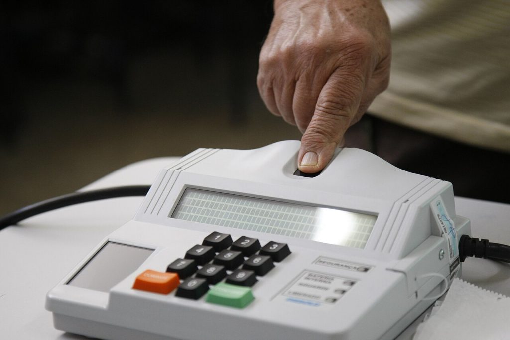 Mão de homem com dedo indicador sobre área de coleta de biometria de uma urna eletrônica.