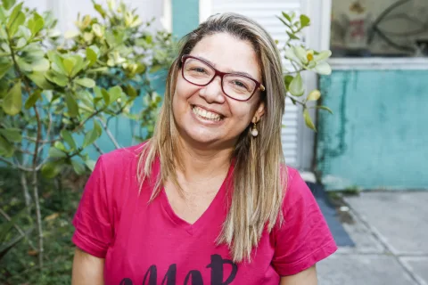 Na foto, mulher jovem sorri em área externa de uma casa.
