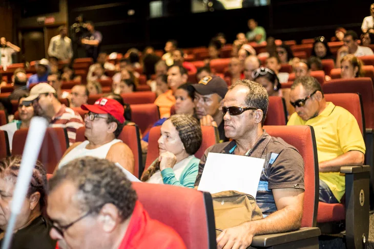 Na foto, várias pessoas sentadas numa sala de cinema com homem de óculos escuros em primeiro plano.