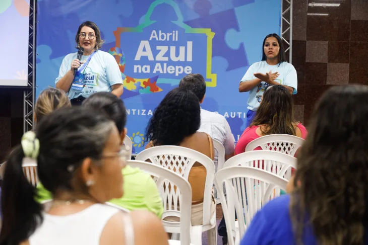 Mulher fala ao microfone com intérprete de Libras ao lado. Elas estão em frente a um painel com fundo azul em que está escrito Abril Azul na Alepe. Pessoas sentadas assistem a palestra.