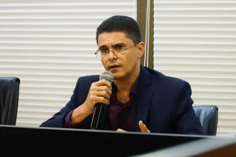 Homem pardo, de óculos e cabelo curto, utilizando blazer azul marinho sobre uma camisa vinho, fala ao microfone