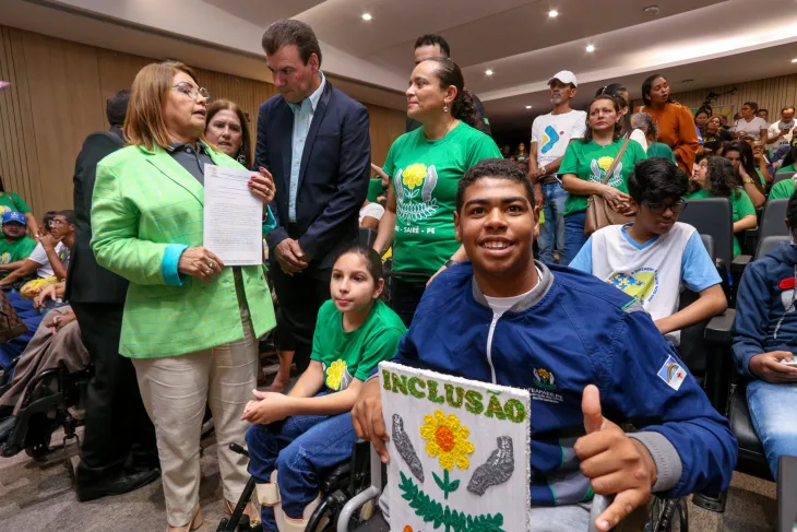 Rapaz em cadeira de rodas segura um cartaz escrito "Inclusão" e com o desenho de um girassol entre duas mãos que apontam para o alto. Ele sorri e faz um símbolo de legal com a outra mão. Ao fundo, em um auditório grande, há várias pessoas vestidas de verde em pé e sentadas.