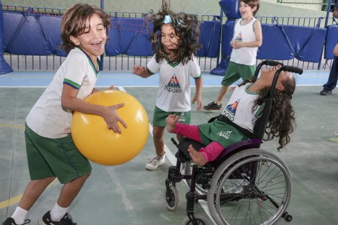 Foto de três crianças jogando com uma bola grande na quadra de uma escola. Uma delas está na cadeira de rodas e ri com a cabeça inclinada para trás.