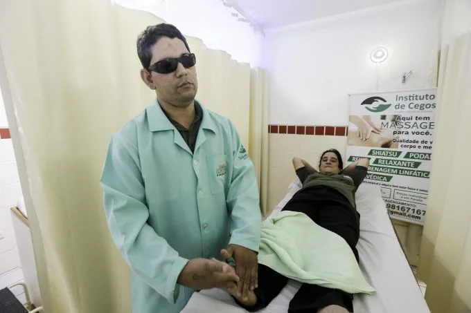 Na foto, homem cego massageia pé de uma mulher deitada na maca.