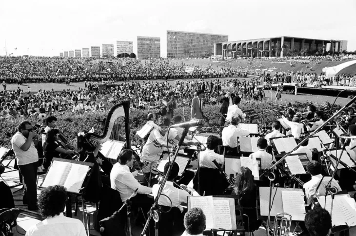 Foto de apresentação da Orquestra Sinfônica (primeiro plano) para milhares de pessoas no gramado do Congresso.