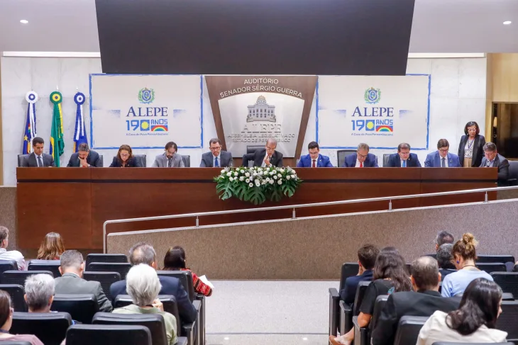 Mesa do auditório Sérgio Guerra, com todas as cadeiras preenchidas por deputados estaduais usando terno e gravata. Atrás dos deputados, do lado esquerdo, estão as bandeiras de Pernambuco, do Brasil e do Recife, penduradas na parede. Ao centro, o nome "Auditório Sérgio Guerra" e um emblema da Alepe com o Palácio Joaquim Nabuco estilizado. Dos dois lados de emblema, o logotipo "Alepe 190 Anos". À frente da mesa onde estão os deputados, as primeiras fileiras do auditórios, parcialmente preenchidas pelo público.